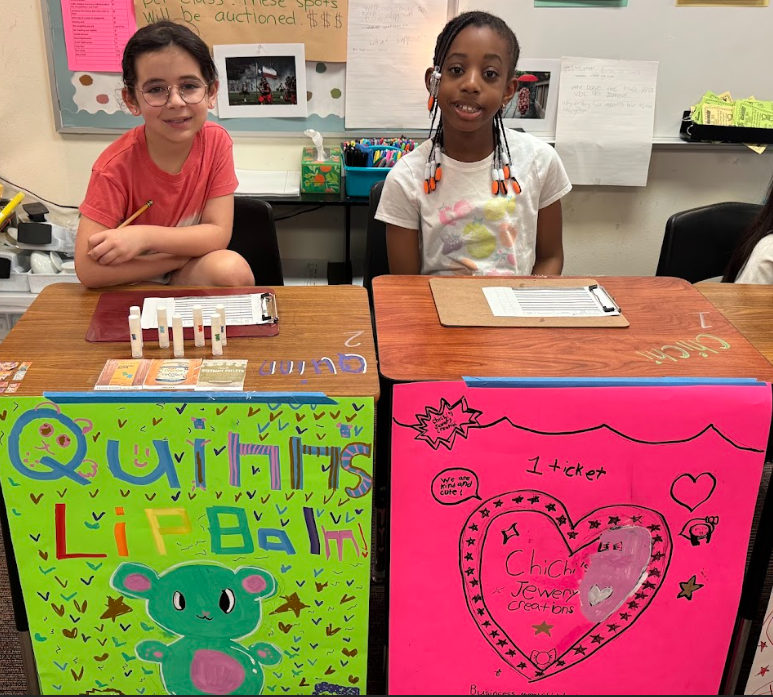 Two students sitting behind desks with signs, selling items for the 3rd Grade Economic Fair