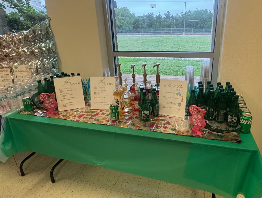 Photograph of table with green table cloth with three drink signs, and various sparkling water, sprite, and syrups, and cups in front of a window