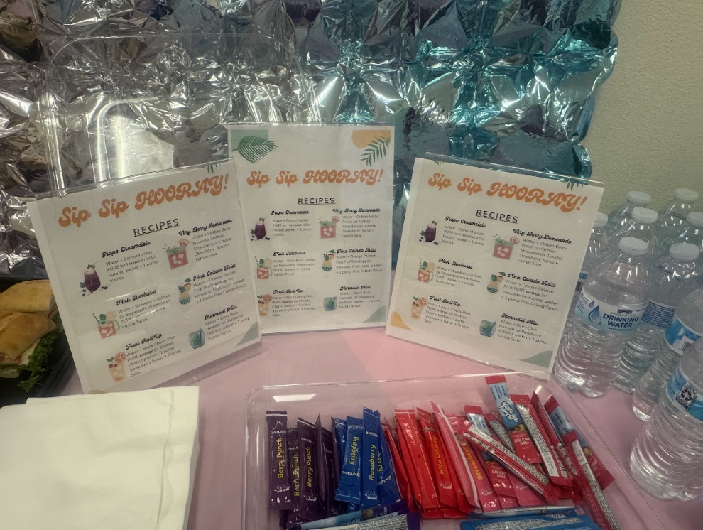 Photograph of table with pink tablecloth, three signs that say "Sip Sip Horray" with drink recipes. There are waters on the right side, individual drink packets in the middle and napkins on the left. 