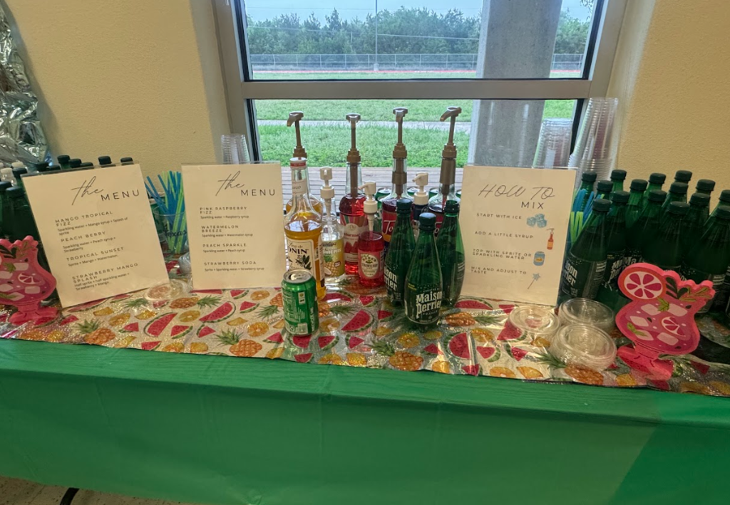 Photograph of table with green table cloth with three drink signs, and various sparkling water, sprite, and syrups, and cups in front of a window