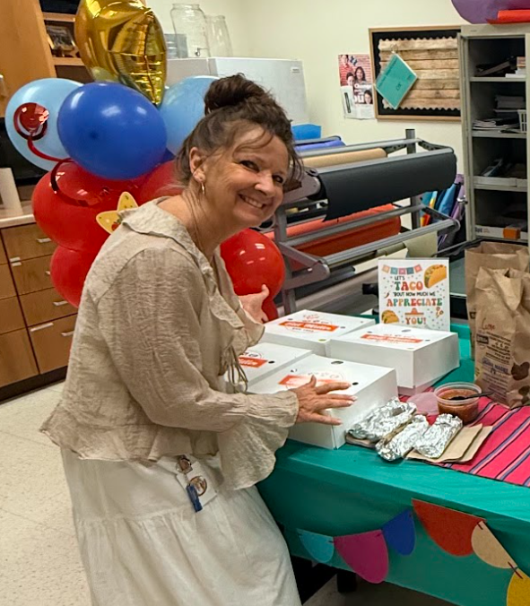 Photograph of teacher standing next to table with 4 white boxes and breakfast tacos wrapped in foil next to balloons