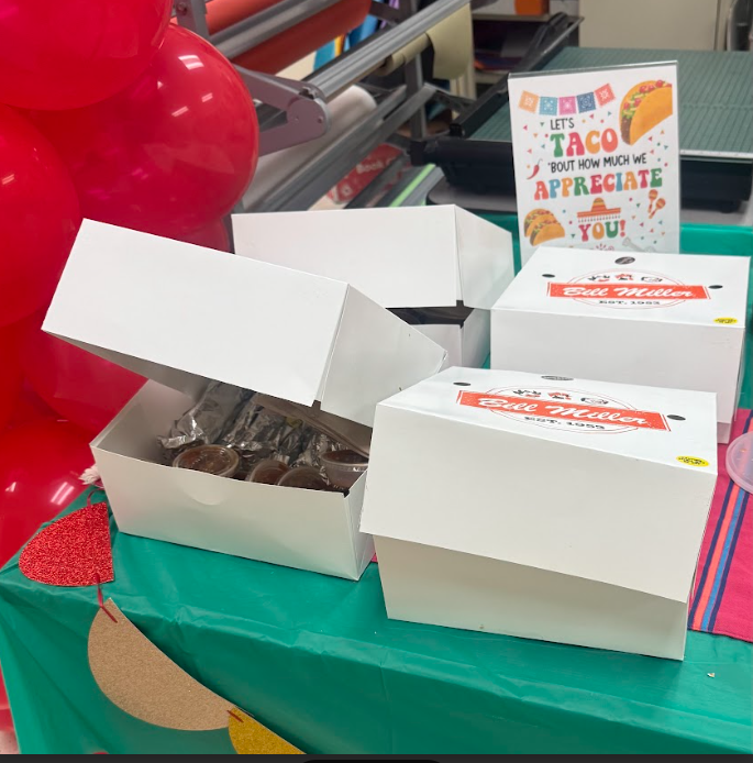 4 white boxes sitting on a table with teal color tablecloth. Sign behind that says "Let's Taco about how much we appreciate you" with a cinco de mayo theme