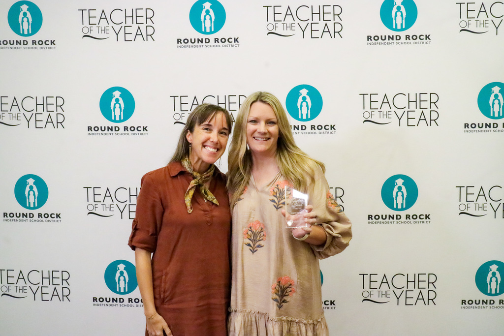 Assitant Principal and Teacher of the Year standing in front of white backdrop that has the Round Rock ISD logo and words "Teacher of the Year"