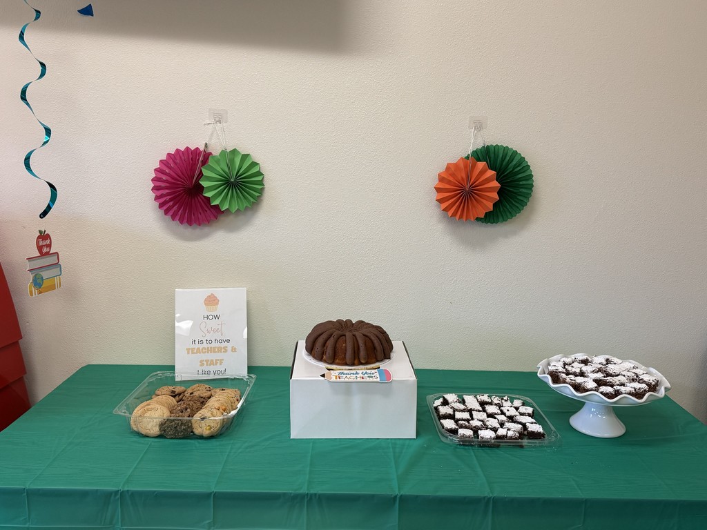 Cookies in container, bundt cake on top of box, a tray of brownies, and a pedastal dish with brownies, sitting on a table with a green tablecloth in front of wall with colorful fan decor. A sign on the table says "How Sweet it is to have Teahcers & Staff Like you!