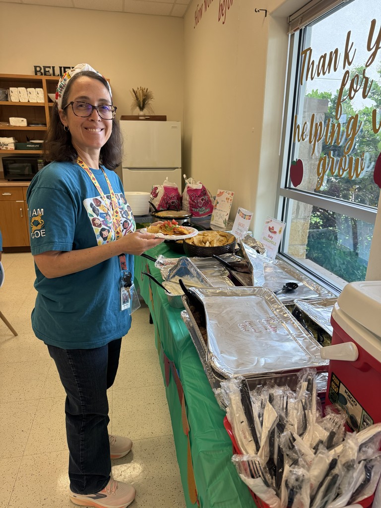 Teacher standing next to table with food, holding a plate of food. 
