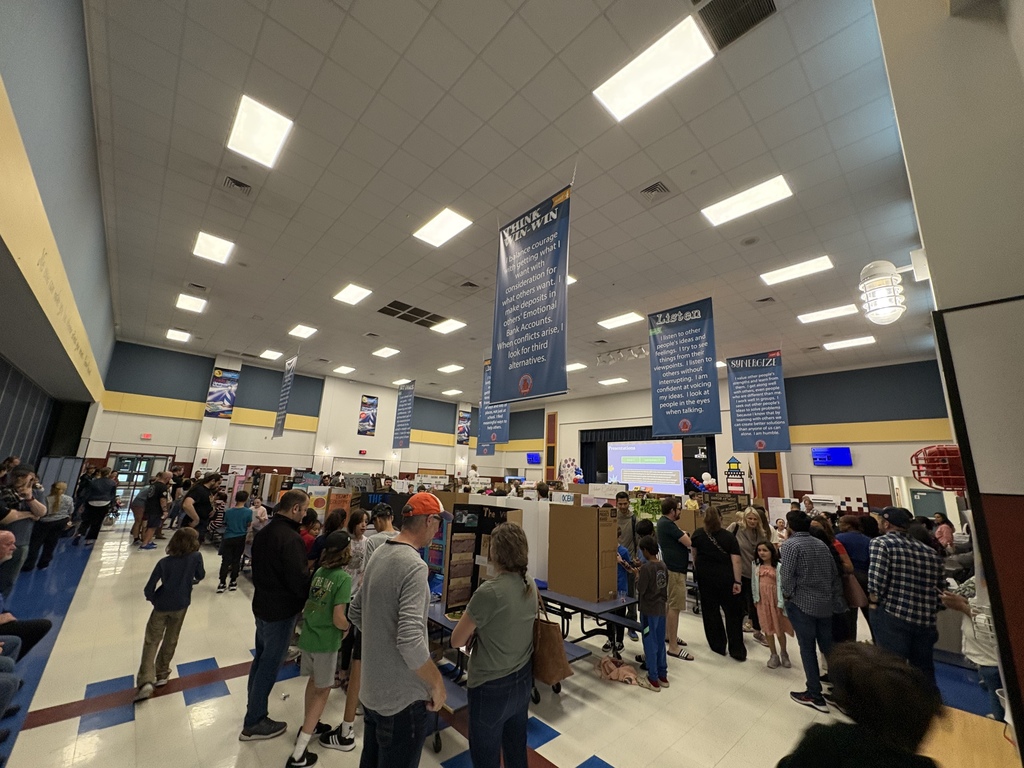 A large, crowded elementary school cafeteria filled with students and parents during a project fair. Blue banners with leadership principles hang from the ceiling over rows of display boards.