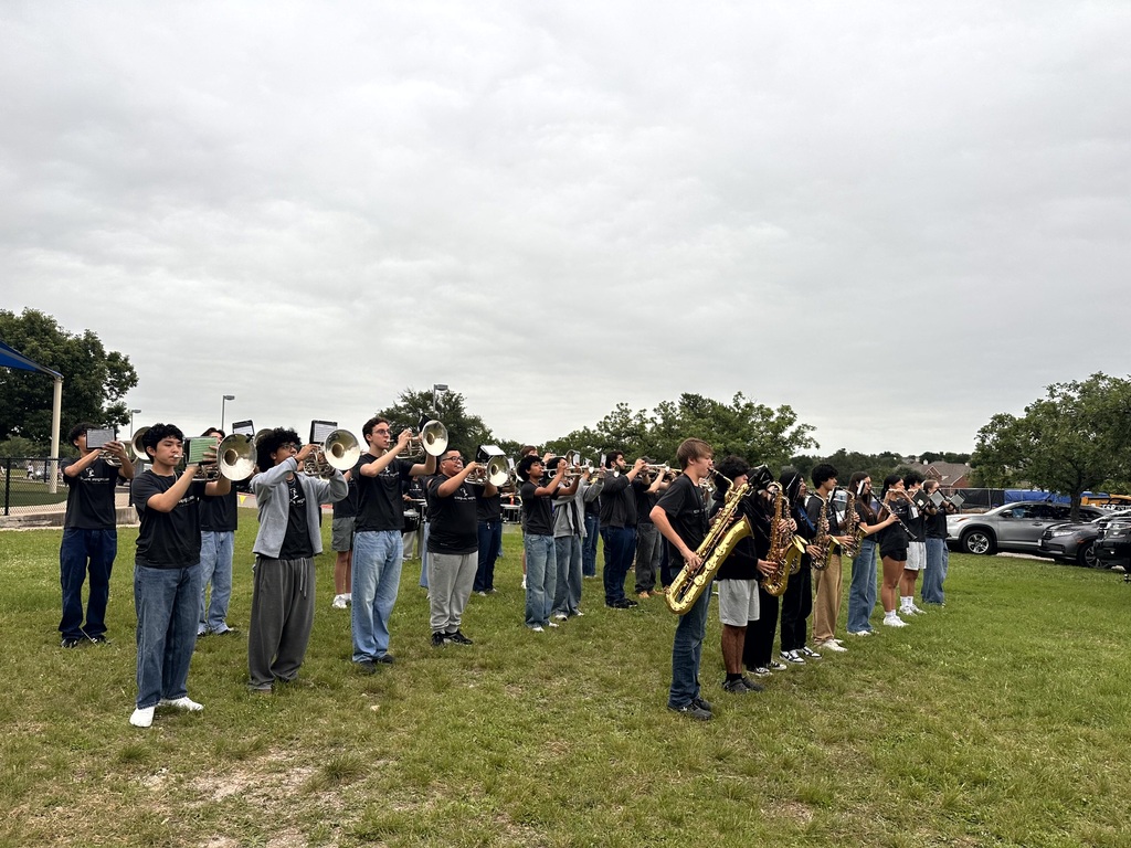 high school marching band in black shirts and jeans performs outdoors on a grass field for an audience.