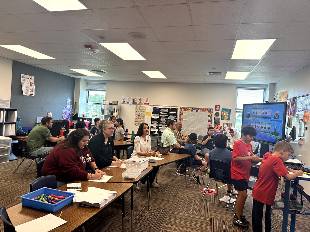 Adults and students sit at classroom tables reviewing leadership binders and watching a digital presentation.