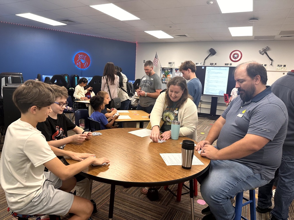 Students and parents play card games together at round tables in a classroom decorated with superhero logos.