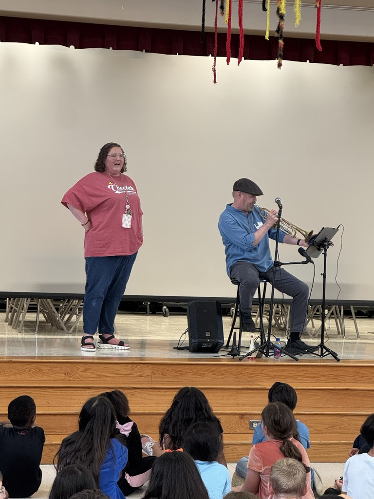 Woman standing on stage next to a person sitting on a stool on a stage, in front of a white screen. He is sittingbehind a microphone and is playing the trumpet in front of a stand in front of students sitting on the floor