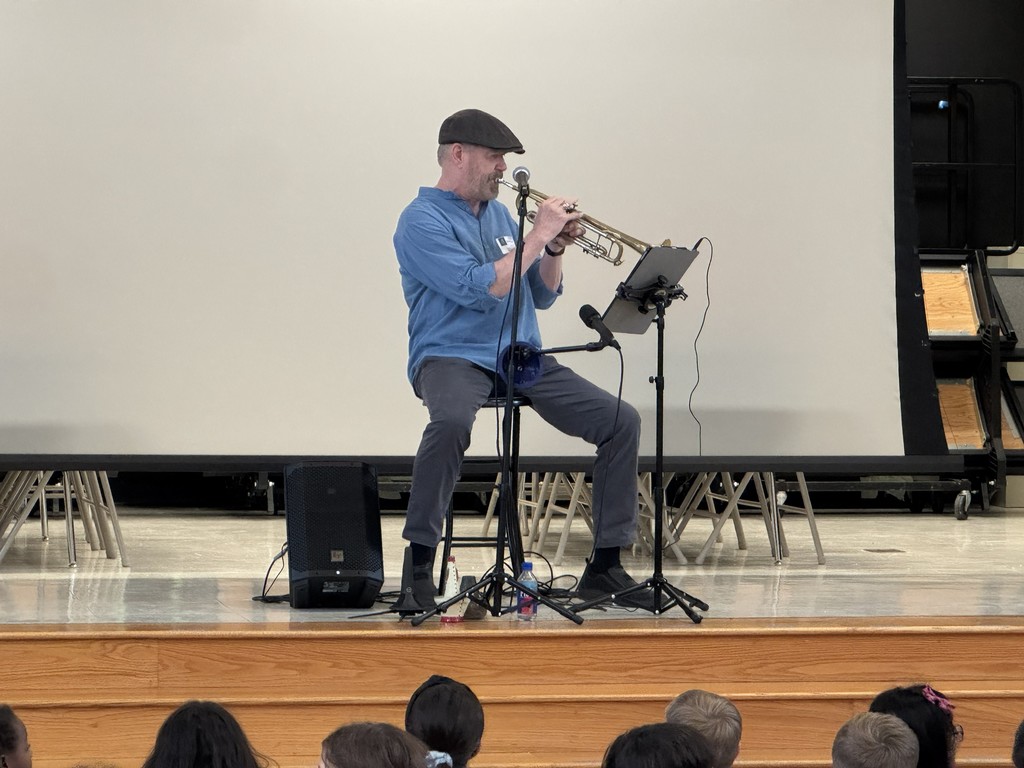 Person sitting on a stool on a stage, in front of a white screen. He is sittingbehind a microphone and is playing the trumpet in front of a stand.