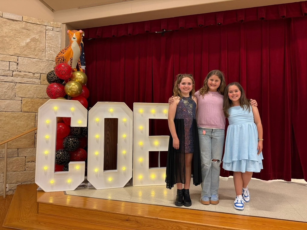Photograph of three girsl standing next to large lit up letters COE in front of red curtain
