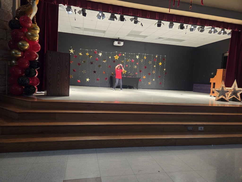 Photograph of student standing on stage in front of hanging stars backdrop. Balloon tower is on the left side of the stage and two stars on the right side of the stage.