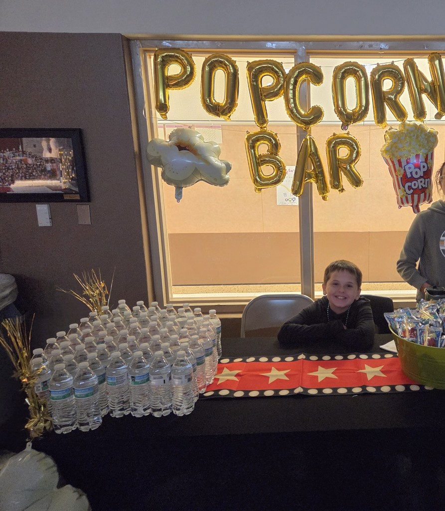 Photograph of student sitting at a table with waters on it in front of a window with balloons that say "Popcorn Bar"