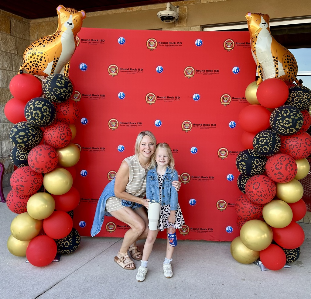 Photograph of and adult and child standing in front of red backdrop with teh Chandler Oaks Logo and IB log on it with the words Round Rock ISD Chandler Oaks Elementary School World IB School. There is a red, black, and gold balloon tower onboth sides of the backdrop with a Cheetah Balloon on top of each