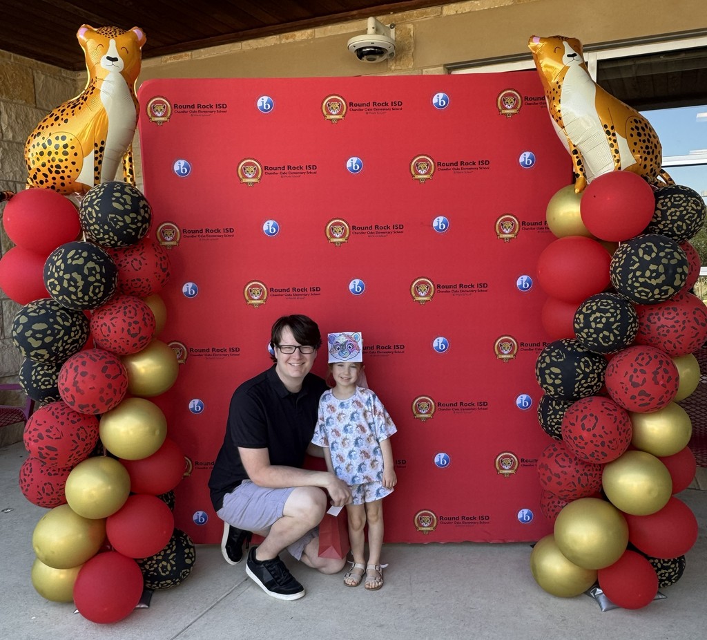 Photograph of an adult and child standing in front of red backdrop with teh Chandler Oaks Logo and IB log on it with the words Round Rock ISD Chandler Oaks Elementary School World IB School. There is a red, black, and gold balloon tower onboth sides of the backdrop with a Cheetah Balloon on top of each