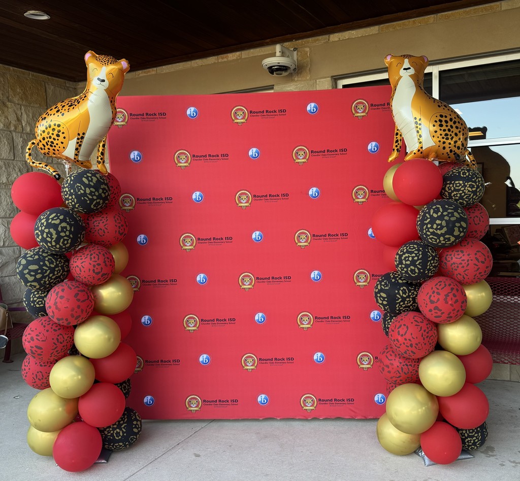 Photograph of red backdrop with teh Chandler Oaks Logo and IB log on it with the words Round Rock ISD Chandler Oaks Elementary School World IB School. There is a red, black, and gold balloon tower onboth sides of the backdrop with a Cheetah Balloon on top of each