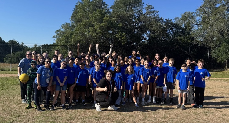 A large group of elementary students in blue shirts and staff members posing together on a grassy field during a sunny day for the annual kickball game.