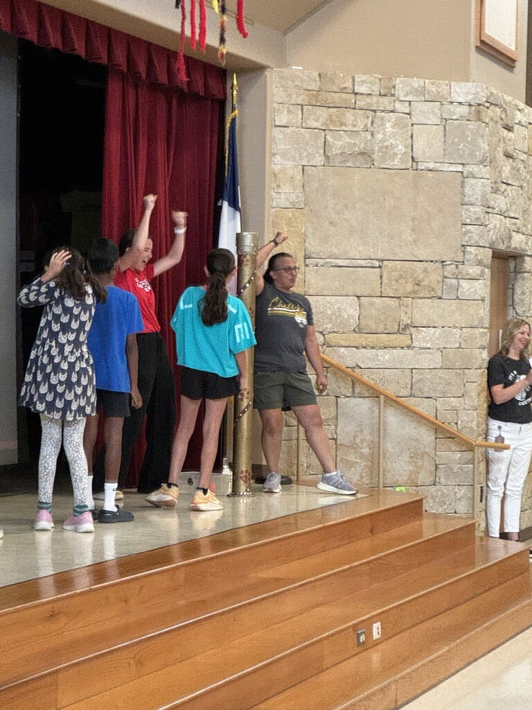 Students and teachers standing on a stage with a red curtain fand flag behind them- and a stone wall to the right of the stage.