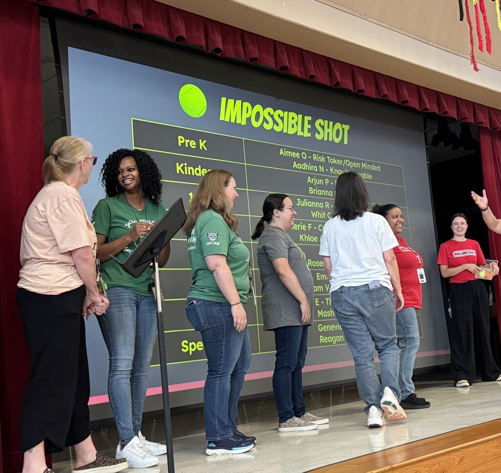 Seven teachers standing in front of a screen that says "Impossible Shot" waiting to try to make the impossible shot
