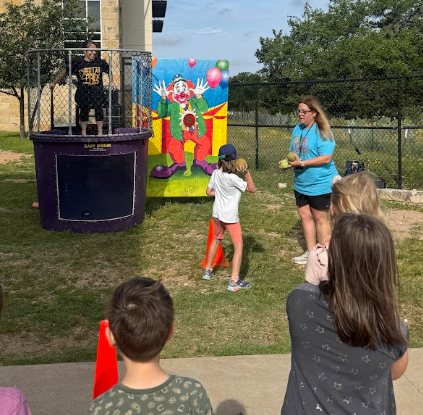 Students watching another student throw a ball a teacher sitting at the top of a dunk tank outside