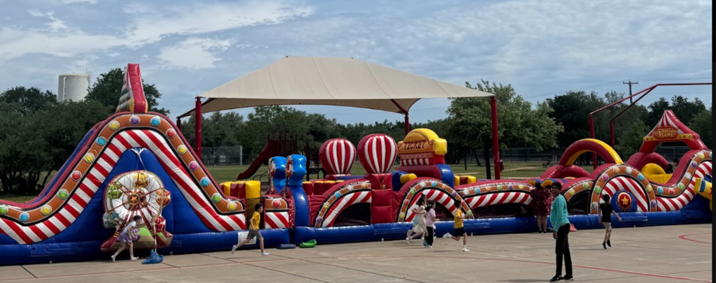 Photograph of large inflatable playscape maze that is carnival themed with students running in front of it and a teacher standing in front of it.