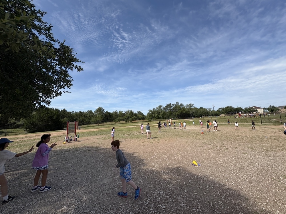 Elementary students and high school athletes play an outdoor game near a shaded school building under a bright sky.