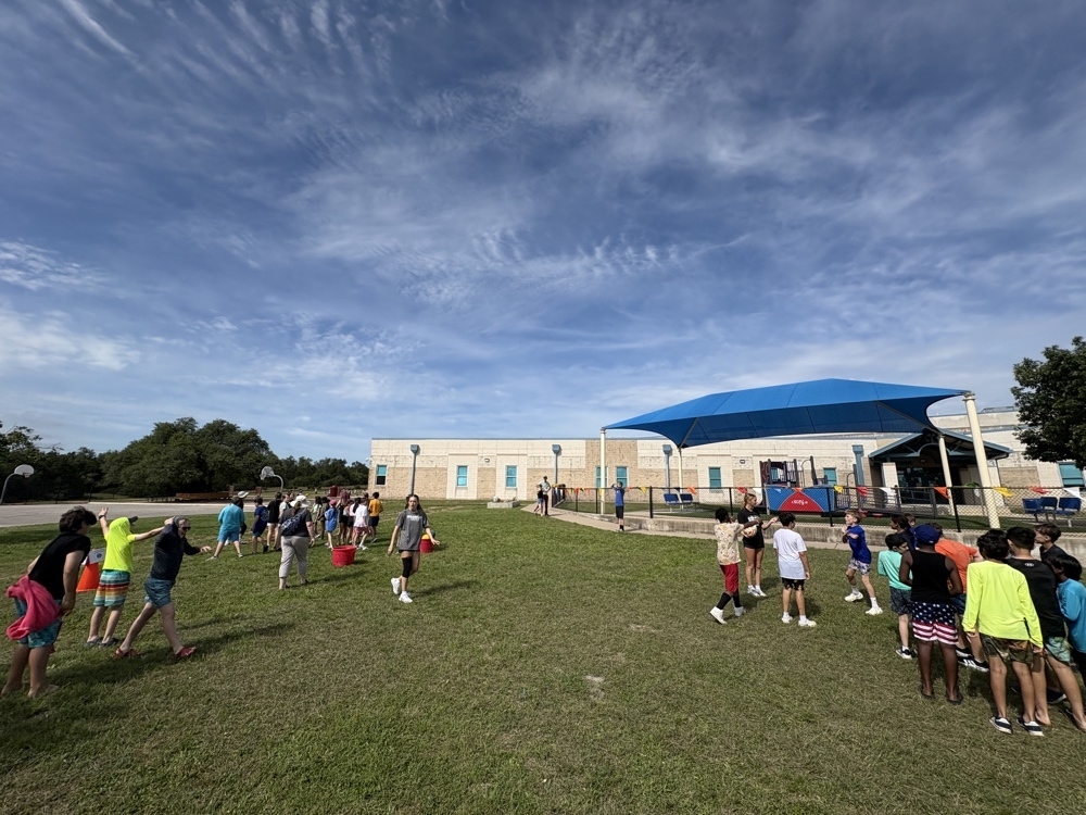 Elementary students and high school athletes play an outdoor game near a shaded school building under a bright sky.