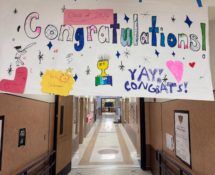 Photograph of a white sign hanging in front of a hallway that says "Class of 2026 Congratulations! with stars, a drawing of a person wearing a graduation cap, pink heart, and a trophy in the middle. Also on the sign, there is a red heart says "Always a Cheetah" and Yellow cloud says "Hello Summer" with a sun