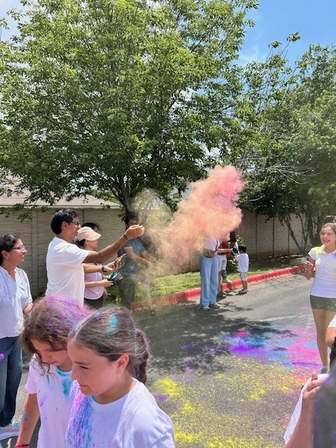 Adult throwing orange/pink dye inot the air next to students walking covered in different color dyes