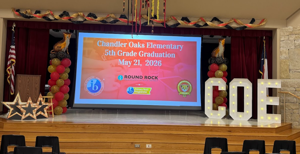 Photograph of a screen that is displaying a red background with the words "Chandler Oaks Elementary 5th Grade Graduation May 21, 2026. On the stage there are two red, yellow, and black balloon towers with a Cheetah Balloon on top, the big lit up letters COE on the right and lit up starts on the left