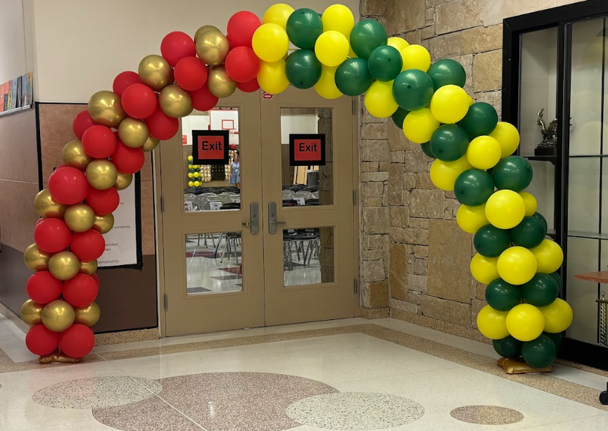 Photograph of a balloon arch in front of the cafeteria doors. On the left side there are red and gold balloons and on the right side there are green and yellow balloons