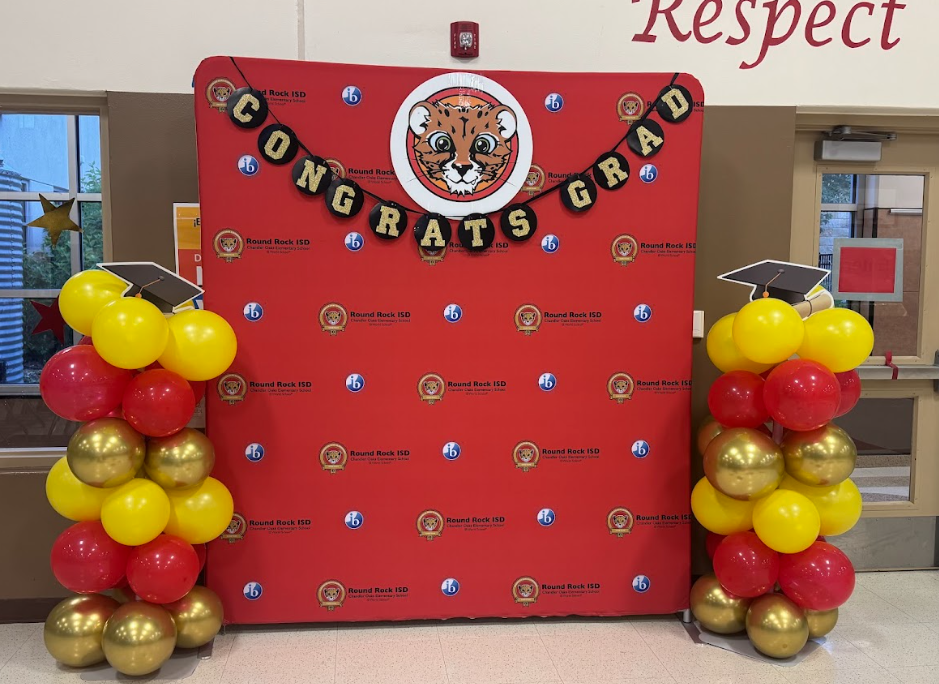 Red photo backdrop with the Chandler Oaks and IB logos on it with a "Congrats Grad" banner on the top and a circle with a cheetah image. On both sides there is a gold, red, and yellow balloon tower with a gradeuation hat on top