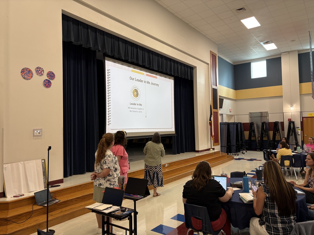 Three presenters stand near the stage in front of a screen displaying a slide titled Our Leader in Me Journey during staff training.