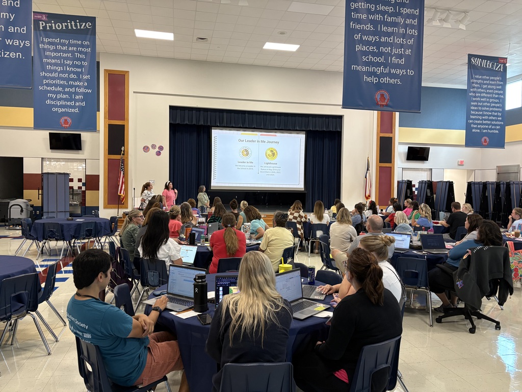 Wide view of BPE staff attending professional development at round tables with laptops under Leader in Me posters in the cafeteria.