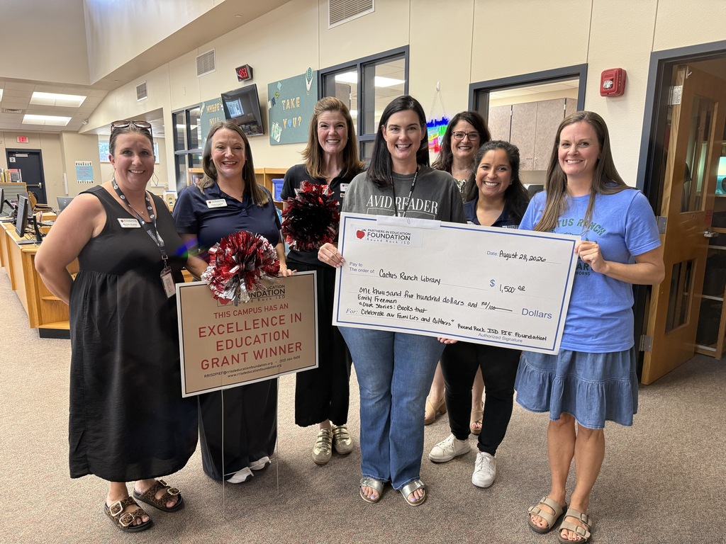 Six smiling Cactus Ranch Elementary staff members stand together in the school library, celebrating a PIE Foundation Excellence in Education Grant. Two women hold a large ceremonial check for $1,500 awarded to the Cactus Ranch Library for books celebrating families and cultures, while another displays a sign announcing the campus as a grant winner.
