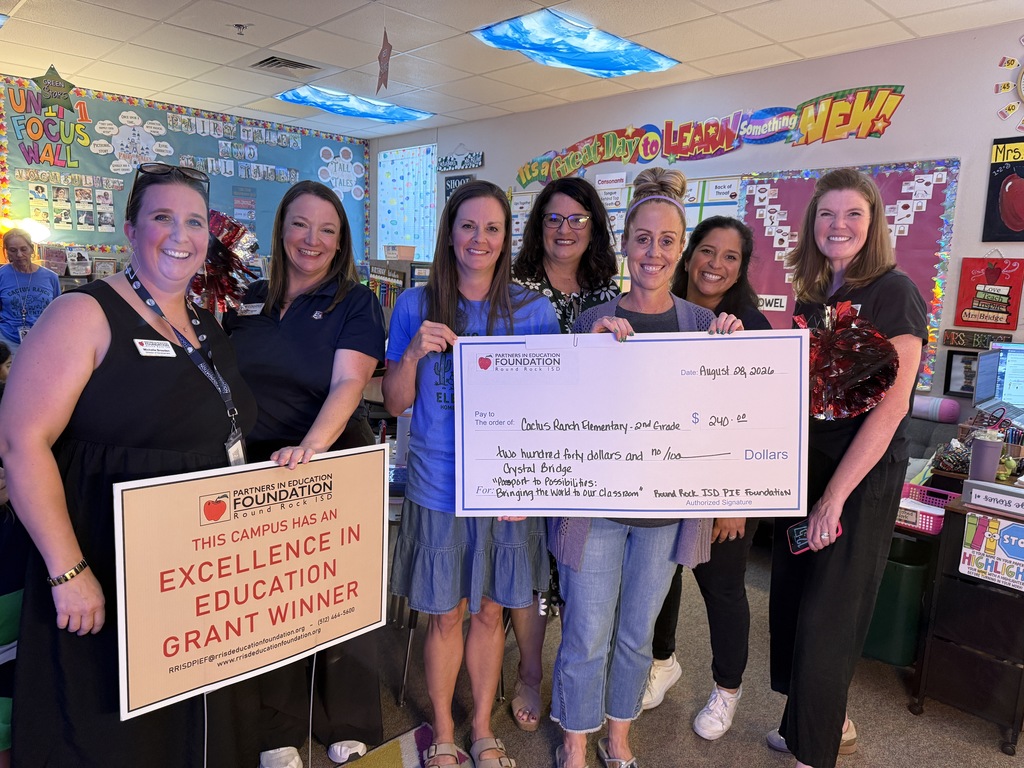 Six smiling Cactus Ranch Elementary staff members stand together in a colorful 2nd grade classroom to celebrate a PIE Foundation Excellence in Education Grant. One woman holds a large ceremonial check for $240 to support virtual field trips that will bring learning experiences from around the world into the classroom, while another displays a sign recognizing the campus as a grant winner.