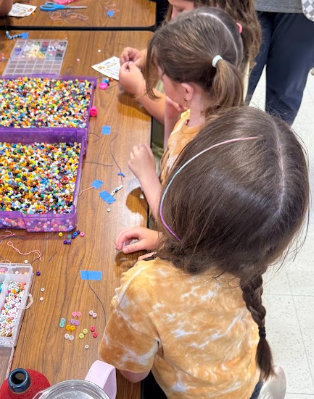 Students sitting at table making bracelets with two bins of beads on the table