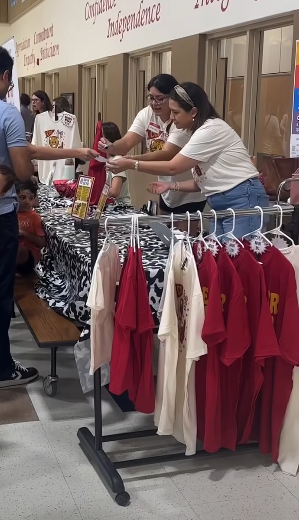 Two adults standing in front of table next to a rack of school red and whitePTA shirts hanging up.