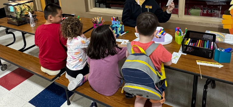 Four students sitting at table using markers to color bookmarks, bins of markers are on the table