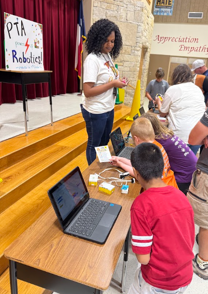 Poster on desk on stage that says "PTA 4th Grade Robotics!". Students looking at chromebooks on desk and lego coding hubs, a teacher who is looking at students working