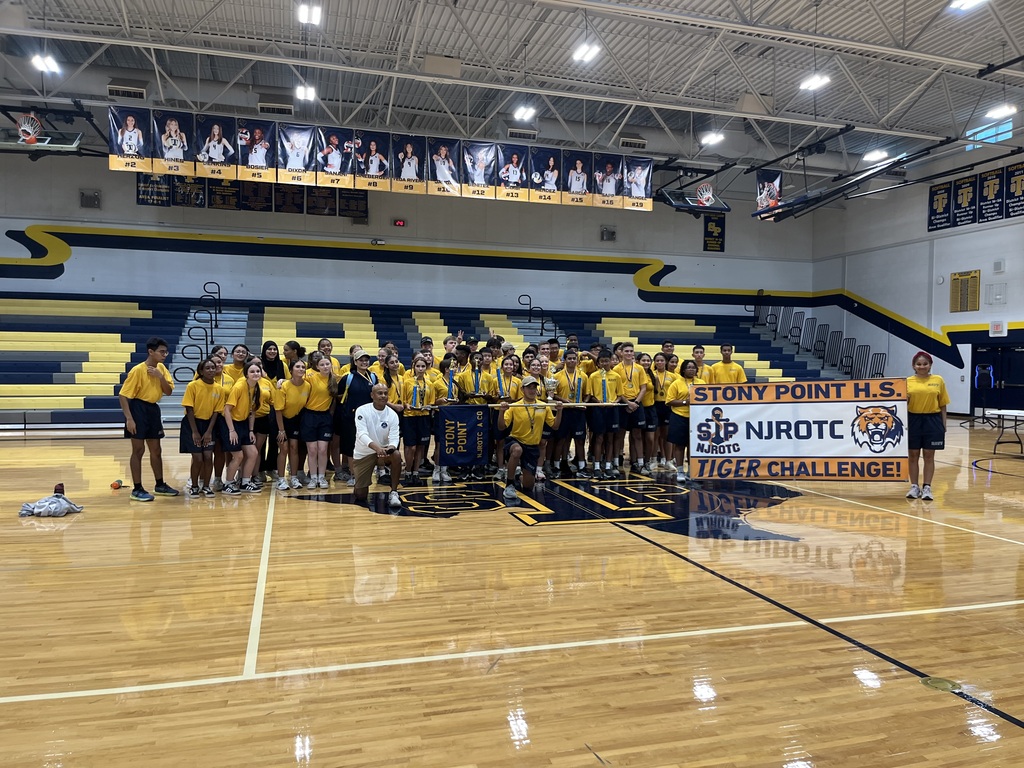 The Stony Point High School NJROTC cadet company and instructors pose together on the gymnasium basketball court following the Tiger Challenge competition. Cadets in yellow and navy polo uniforms proudly hold up a large trophy mounted on poles along with a banner that reads "Stony Point H.S. NJROTC Tiger Challenge!