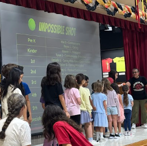 Students are standing in a line facing a teacher next to a screen that ays "Impossible Shot" with grade levels and names