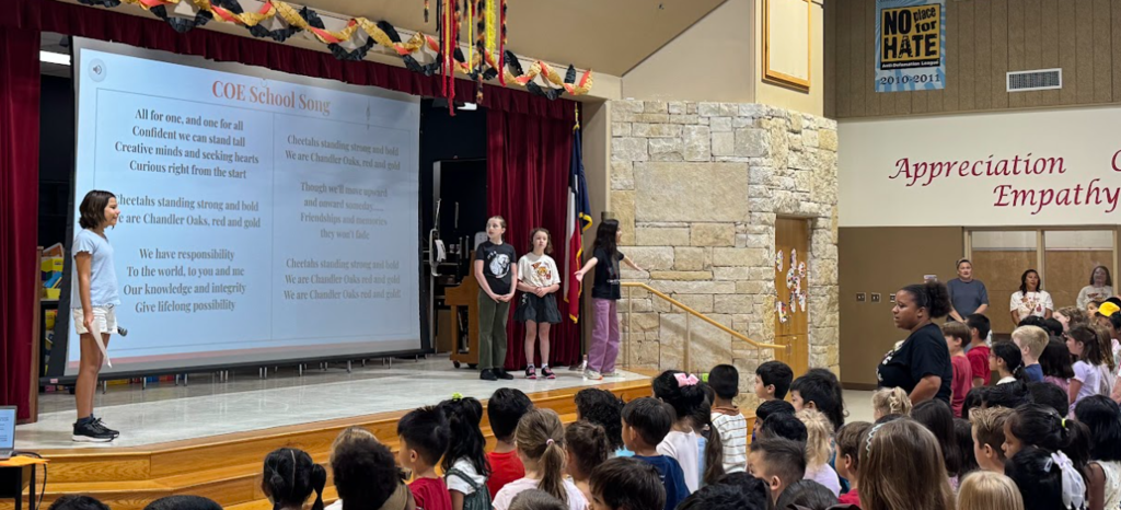 Students standing on stage next to a screen that says "COE School Song" with the lyrics. Students and teachers are standing up facing the screen and stage 