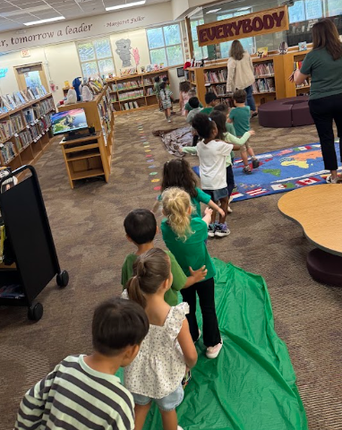 A line of young children seen from behind, following each other along a long green plastic runner laid out across the carpeted floor of a library. Bookcases line the background wall beneath a sign that reads "EVERYBODY."