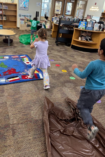 A group of young students wearing casual clothes running through an indoor path setup in a school library. A child in a blue shirt is jumping over a crumpled brown tarp on the carpet, while another child in a light purple dress walks along a path of colorful markers next to a world map rug.