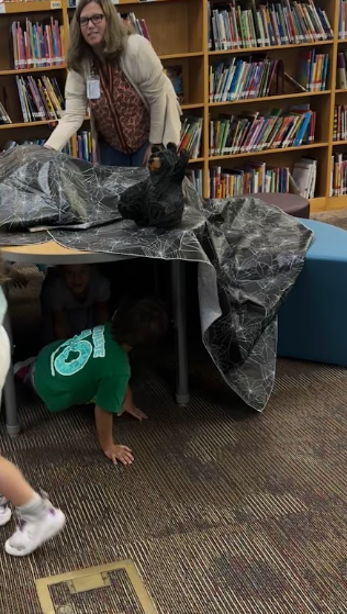 A child wearing a green shirt crawling under a round wooden table draped in dark, spiderweb-patterned fabric and decorated with a bear figure. A female teacher wearing glasses and a cardigan stands behind the table in front of tall wooden bookshelves filled with books.