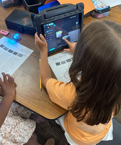 An overhead view focusing on a student with long brown hair, sitting at a desk and using an iPad in a thick protective case. She is dragging and dropping colorful block code on the screen. Next to the iPad is a worksheet titled "Dash's Main Menu."