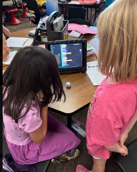 Two students with long hair (one dark, one blonde) seated at a wooden desk in a busy classroom. They are both looking at an iPad showing code, with a small silver Ozobot on the desk between them. Other classroom activities are visible in the background.