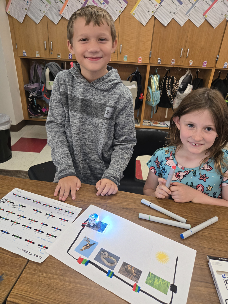 A young boy and girl smiling at the camera, working together at a desk with an Ozobot robot. In front of them is a laminated "Food Chain Color Codes" sheet featuring pictures of a sun, grass, mouse, snake, and owl connected by marker lines. The Ozobot is glowing and navigating the path.